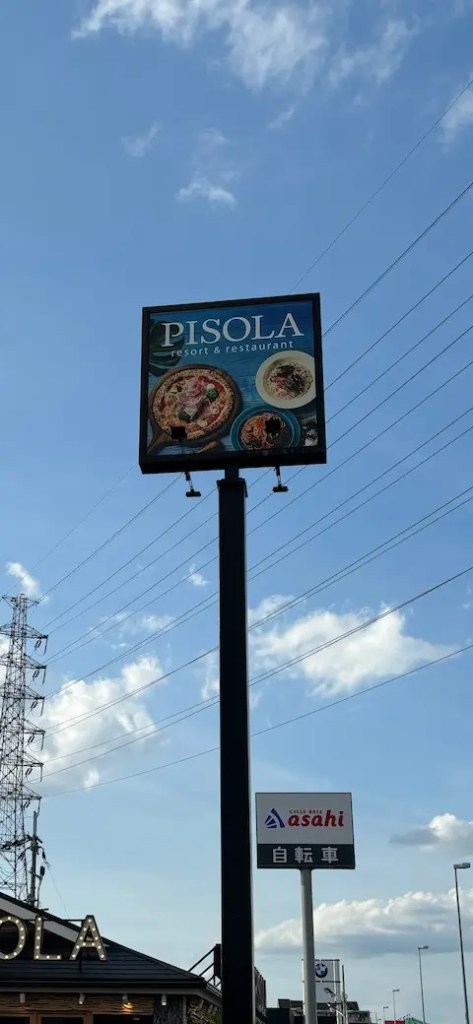 青空の下、レストラン「PISOLA」の看板が立っている。看板には料理の写真が描かれている。