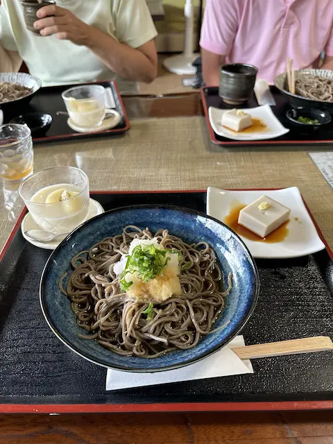 そばが盛られた青い皿と、大根おろしとネギがトッピングされている。テーブルには他の料理や飲み物も見える。