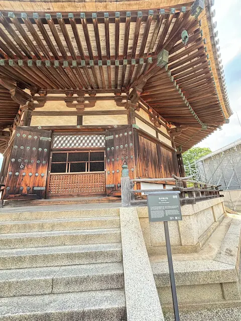 伝統的な日本の寺院の入口で、木製の玄関扉と豪華な屋根が特徴的。階段の前には説明板が設置されている。