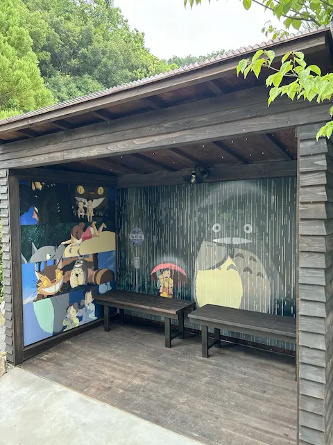 宮崎駿のアニメキャラクターが描かれた小屋の内部、木製のベンチが2つあり、周囲に緑がある
