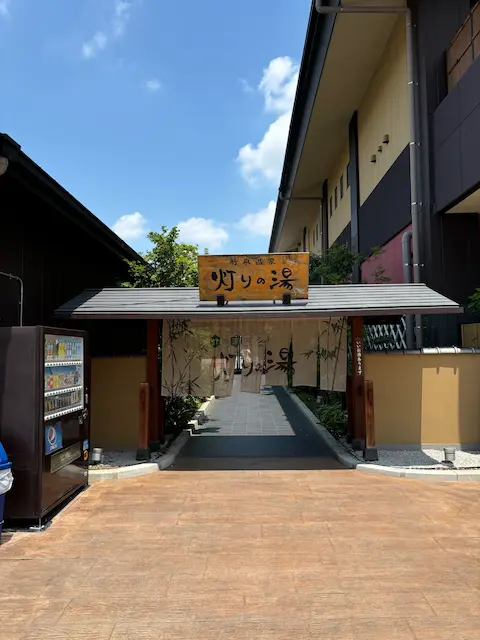 温泉の入り口、灯りの湯の看板が見える。周囲には緑があり、青空が広がっている。
